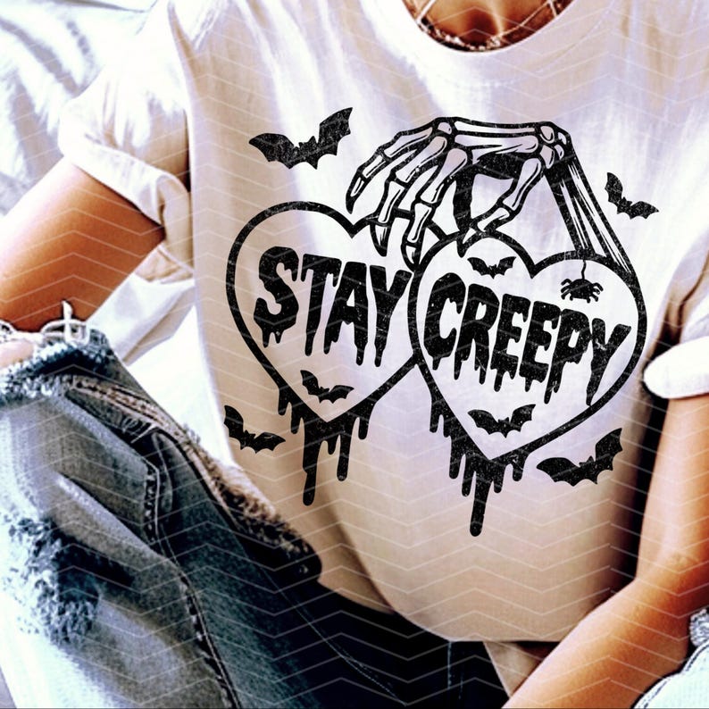 May include: Cream-coloured t-shirt with a black graphic. The design features two heart shapes with the words "STAY CREEPY", held by a skeleton hand. Bats and dripping details add to the spooky aesthetic.