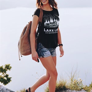 May include: A woman wearing a straw hat, sunglasses, and a black t-shirt with a white graphic and the text "LAKE LIFE CUZ BEACHES BE SALTY". She is wearing denim shorts, sandals, and a brown backpack.