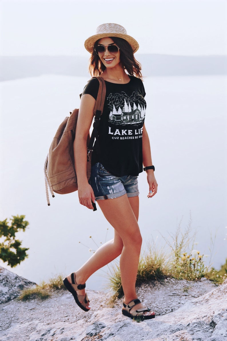 May include: A woman wearing a straw hat, sunglasses, and a black t-shirt with a white graphic and the text "LAKE LIFE CUZ BEACHES BE SALTY". She is wearing denim shorts, sandals, and a brown backpack.