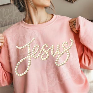 Puede incluir: Una sudadera rosa claro con la palabra "Jesus" escrita con cuentas de perlas falsas. La sudadera tiene cuello redondo y mangas largas. El nombre de la marca "Magic Mango" est&aacute; impreso en la prenda.