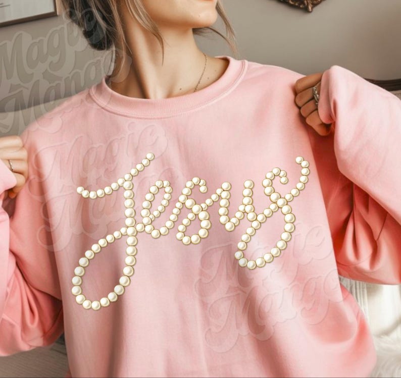 Puede incluir: Una sudadera rosa claro con la palabra "Jesus" escrita con cuentas de perlas falsas. La sudadera tiene cuello redondo y mangas largas. El nombre de la marca "Magic Mango" est&aacute; impreso en la prenda.