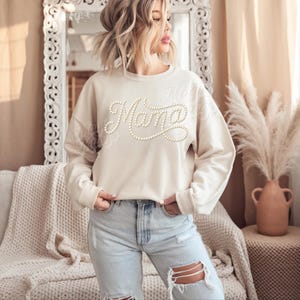 May include: Cream-colored sweatshirt with the word "Mama" spelled out in a cursive font, embellished with small, pearl-like beads. The long-sleeved top is paired with light-wash, ripped jeans. The background includes a mirror, couch, and decorative vase.