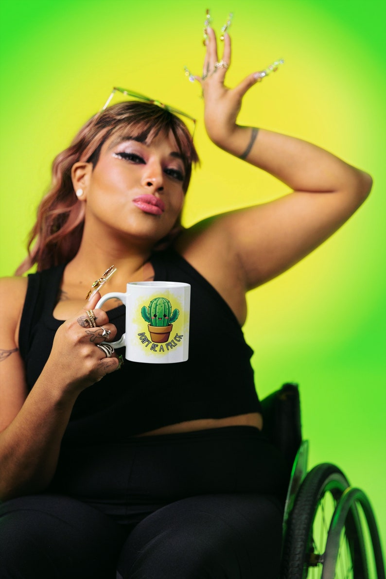May include: A white ceramic mug with a green cactus illustration and the text "Don't be a prick". The mug is being held by a person in a black tank top and black pants.