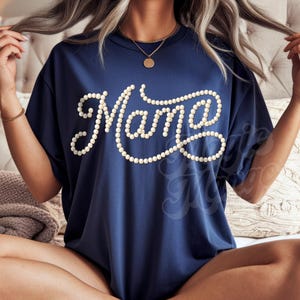 May include: Navy blue t-shirt with the word "Mama" in white, stylized with a pearl-like design. The shirt has short sleeves and a round neckline. The shirt is worn by a person sitting on a bed.
