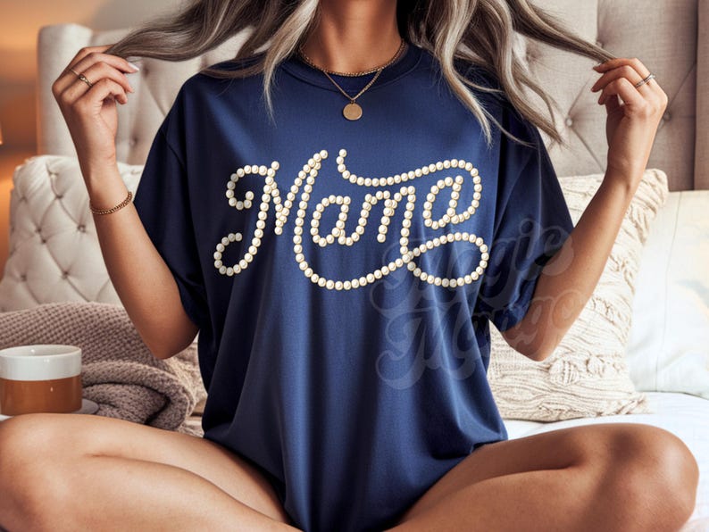 May include: Navy blue t-shirt with the word "Mama" in white, stylized with a pearl-like design. The shirt has short sleeves and a round neckline. The shirt is worn by a person sitting on a bed.