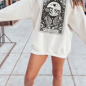 May include: A white sweatshirt featuring a black and white illustration of a tarot card depicting two skeletons holding hands. The card is labelled "The Lovers".