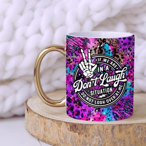 May include: A white ceramic mug with a gold handle. The mug has a colorful design with a peace sign, stars, and the text "If we are in a Don't Laugh situation do not look over at me".