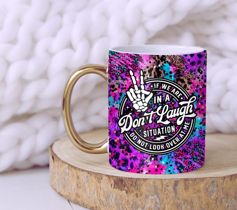 May include: A white ceramic mug with a gold handle. The mug has a colorful design with a peace sign, stars, and the text "If we are in a Don't Laugh situation do not look over at me".