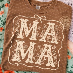 Puede incluir: Una camiseta marr&oacute;n con un dise&ntilde;o floral blanco que dice "Mama" en una fuente de estilo vintage.