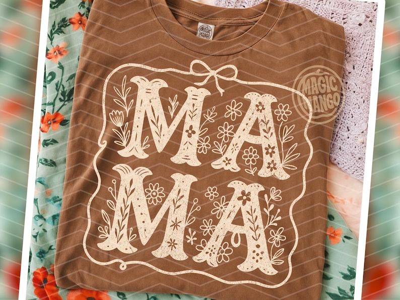 Puede incluir: Una camiseta marr&oacute;n con un dise&ntilde;o floral blanco que dice "Mama" en una fuente de estilo vintage.