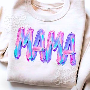 May include: A cream-coloured sweatshirt featuring the word "MAMA" in large, colourful, textured lettering. The letters are painted with shades of pink, blue, and purple, giving a brushstroke effect. The sweatshirt has a crew neck.