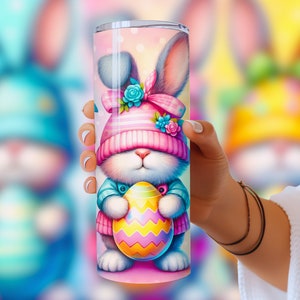 May include: A pink and blue stainless steel tumbler with a cartoon bunny holding a colorful Easter egg. The bunny is wearing a pink and white hat with a blue bow.
