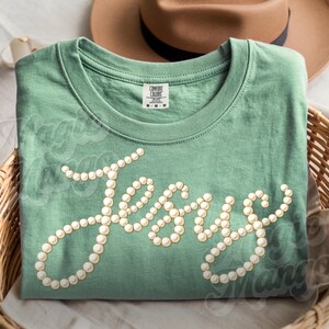 Puede incluir: Camiseta verde salvia con la palabra "Jesus" escrita en cursiva con adornos de perlas. La camiseta est&aacute; doblada en una cesta de mimbre, con un sombrero marr&oacute;n en el fondo.