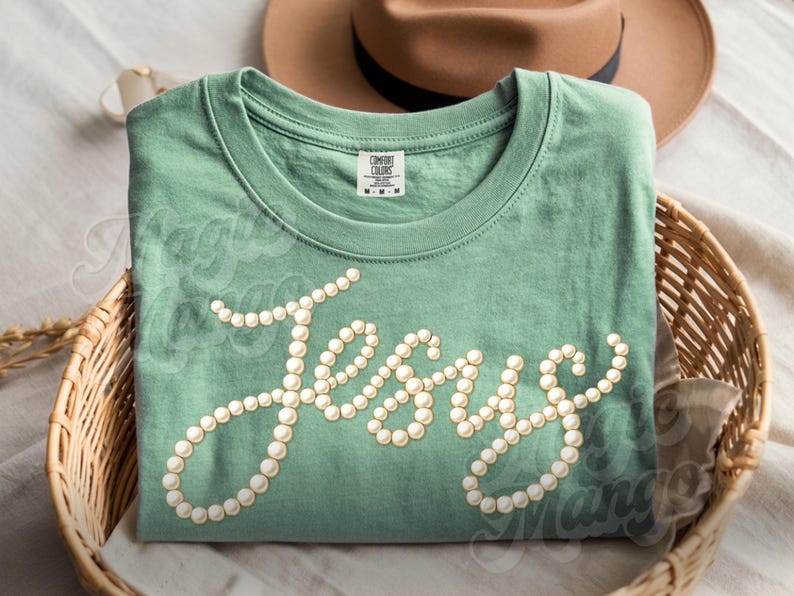 Puede incluir: Camiseta verde salvia con la palabra "Jesus" escrita en cursiva con adornos de perlas. La camiseta est&aacute; doblada en una cesta de mimbre, con un sombrero marr&oacute;n en el fondo.