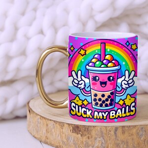 May include: A white ceramic mug with a gold handle features a cartoon illustration of a pink bubble tea cup with a rainbow and stars. The text "SUCK MY BALLS" is printed on the mug.