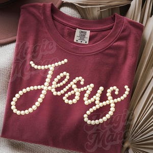 Puede incluir: Camiseta granate con la palabra "Jesus" escrita con perlas. Las palabras "Magic Man" son ligeramente visibles en el fondo. La camiseta est&aacute; doblada y se muestra con hojas de palma secas.