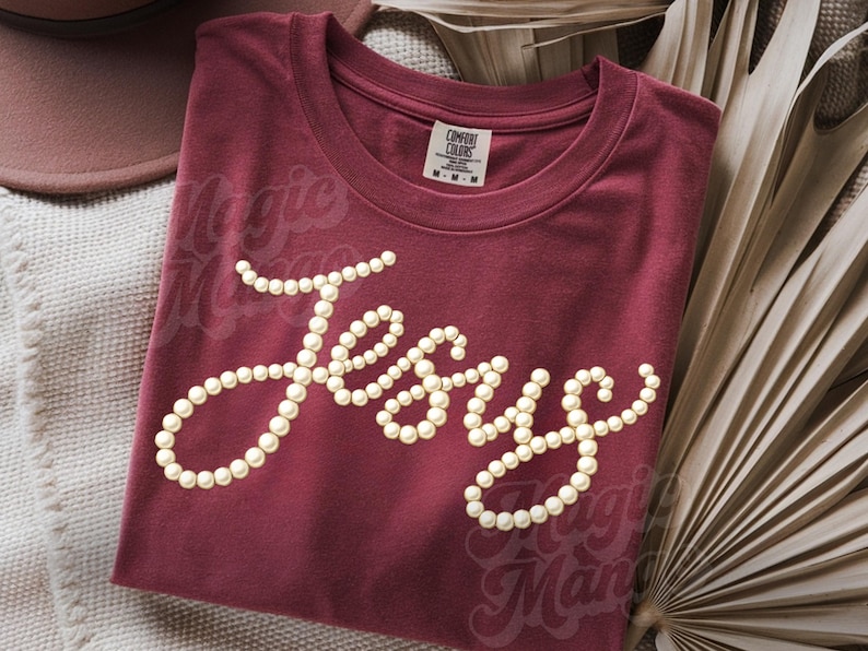 Puede incluir: Camiseta granate con la palabra "Jesus" escrita con perlas. Las palabras "Magic Man" son ligeramente visibles en el fondo. La camiseta est&aacute; doblada y se muestra con hojas de palma secas.