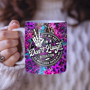 May include: A white ceramic mug with a pink, purple, and blue abstract design. The mug features a black and white graphic of a hand with a peace sign and the text "If we are in a situation don't laugh do not look over at me".
