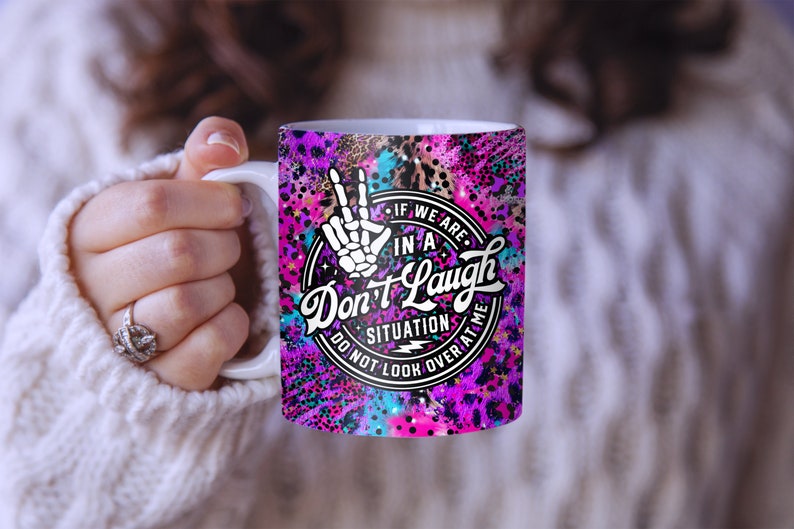 May include: A white ceramic mug with a pink, purple, and blue abstract design. The mug features a black and white graphic of a hand with a peace sign and the text "If we are in a situation don't laugh do not look over at me".