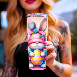 May include: A pink and blue Easter bunny with a pink and white striped hat holds a yellow and pink Easter egg. The bunny is on a pink background with white polka dots. The image is on a stainless steel tumbler.