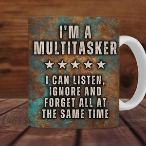May include: A white ceramic coffee mug with a brown, blue, and green design. The mug has the text "I'M A MULTITASKER" with five stars and the text "I CAN LISTEN, IGNORE AND FORGET ALL AT THE SAME TIME".