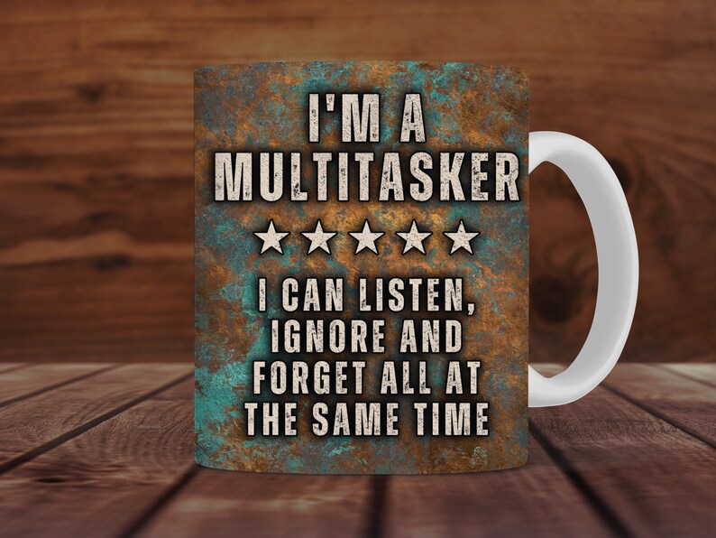 May include: A white ceramic coffee mug with a brown, blue, and green design. The mug has the text "I'M A MULTITASKER" with five stars and the text "I CAN LISTEN, IGNORE AND FORGET ALL AT THE SAME TIME".