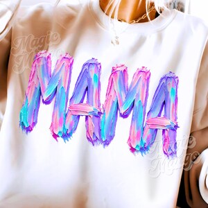 May include: Cream-coloured sweatshirt featuring the word "MAMA" in large, textured brushstroke lettering. The letters are a blend of pink, blue, and purple, creating a vibrant, artistic design. A casual, comfortable top.