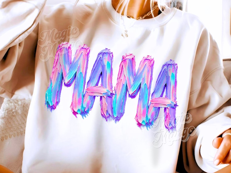 May include: Cream-coloured sweatshirt featuring the word "MAMA" in large, textured brushstroke lettering. The letters are a blend of pink, blue, and purple, creating a vibrant, artistic design. A casual, comfortable top.