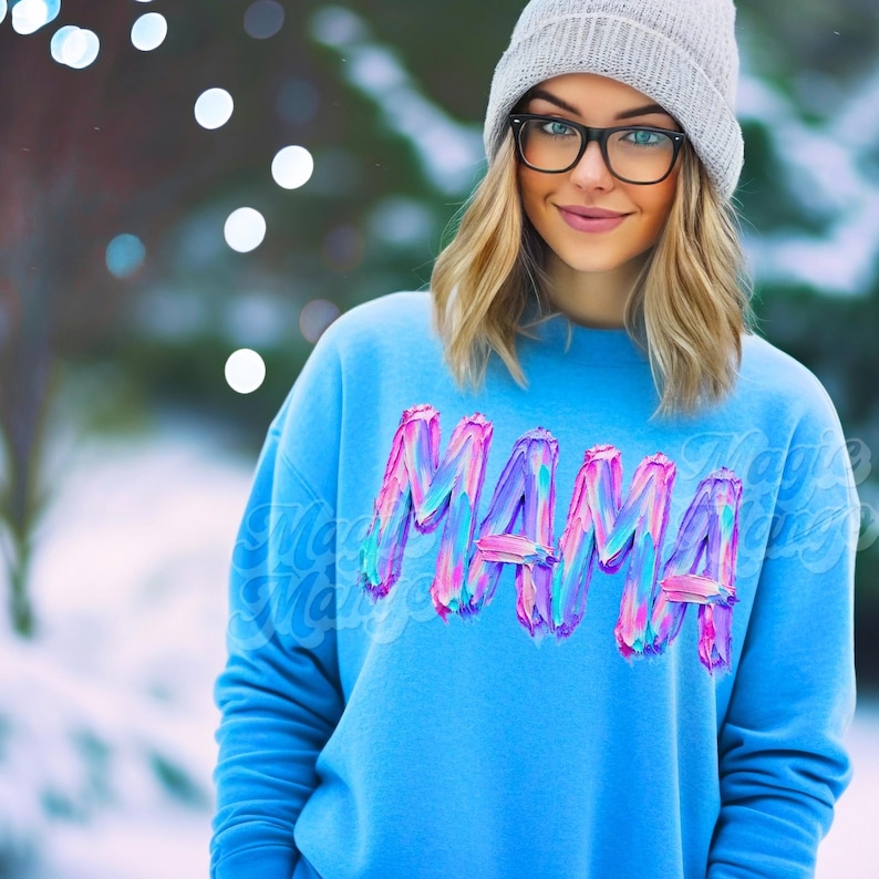 May include: A light blue sweatshirt with the word "MAMA" in a colourful, textured font. The sweatshirt is worn with a grey beanie and glasses. The background is blurred with bokeh lights.