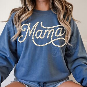 May include: A blue sweatshirt with the word "Mama" in a cursive font, outlined with small, white, pearl-like beads. The sweatshirt has long sleeves and a crew neck.