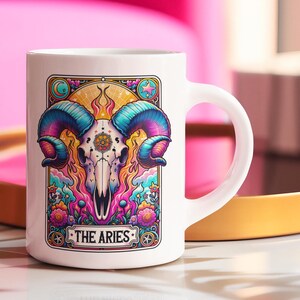 May include: White ceramic mug with a colourful illustration of an Aries ram skull, featuring blue, purple, and yellow accents. The design includes floral elements and the text "THE ARIES". A handle is visible on the right side.