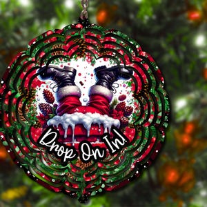 May include: A red and green Christmas wind spinner with a Santa Claus design. Santa is upside down with his legs sticking out of a chimney. The text "Drop On In!" is written on the spinner.