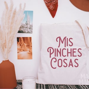 May include: White canvas tote bag with the text "Mis Pinches Cosas" in pink lettering. The bag is sitting on a table with a black and white patterned surface.