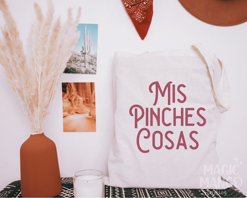 May include: White canvas tote bag with the text "Mis Pinches Cosas" in pink lettering. The bag is sitting on a table with a black and white patterned surface.