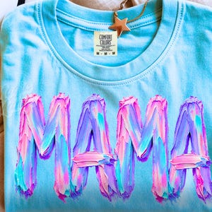 May include: A pale blue t-shirt featuring the word "MAMA" in large, textured lettering. The letters are painted with strokes of pink, purple, and blue. A gold star pendant hangs near the collar.