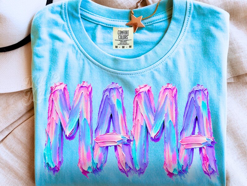 May include: A pale blue t-shirt featuring the word "MAMA" in large, textured lettering. The letters are painted with strokes of pink, purple, and blue. A gold star pendant hangs near the collar.
