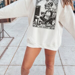 Op de afbeelding: Een wit sweatshirt met een zwart-witte illustratie van een skelet dat videogames speelt. De illustratie is omlijst met een zwarte rand met de tekst "THE GAMER" onderaan.