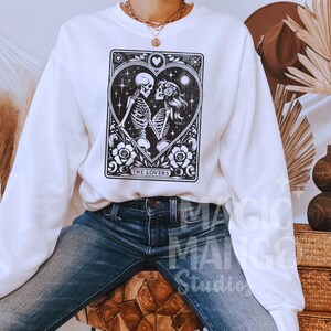 May include: White sweatshirt featuring a black and white tarot card design with two skeletons embracing inside a heart. The card reads "THE LOVERS" at the bottom. The sweatshirt is a crew neck style.