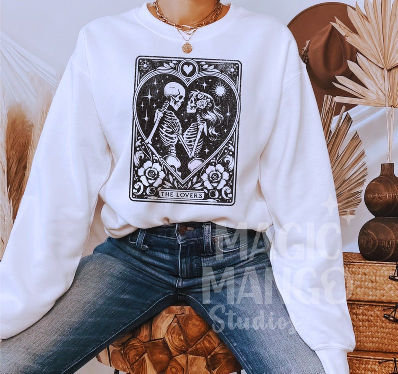 May include: White sweatshirt featuring a black and white tarot card design with two skeletons embracing inside a heart. The card reads "THE LOVERS" at the bottom. The sweatshirt is a crew neck style.