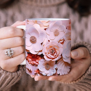 K&ouml;nnte beinhalten: Eine wei&szlig;e Keramiktasse mit einem Blumenmuster. Das Design zeigt rosa, pfirsichfarbene und korallenfarbene Papierblumen.