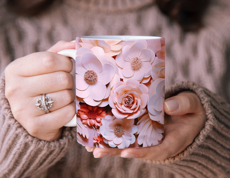 K&ouml;nnte beinhalten: Eine wei&szlig;e Keramiktasse mit einem Blumenmuster. Das Design zeigt rosa, pfirsichfarbene und korallenfarbene Papierblumen.