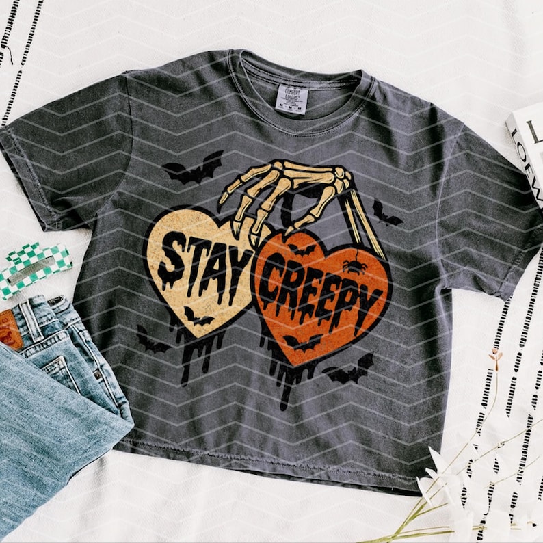 May include: Dark grey t-shirt with a Halloween graphic. The design shows a skeleton hand holding two heart-shaped designs with the words "Stay Creepy". Bats and dripping accents complete the spooky design.