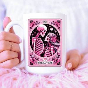 K&ouml;nnte beinhalten: Wei&szlig;e Keramik-Tasse mit einer rosa-schwarzen Illustration von zwei sich umarmenden Skeletten, mit den Worten "AMOR" und "THE LOVERS". Das Design enth&auml;lt florale und sternf&ouml;rmige Elemente.