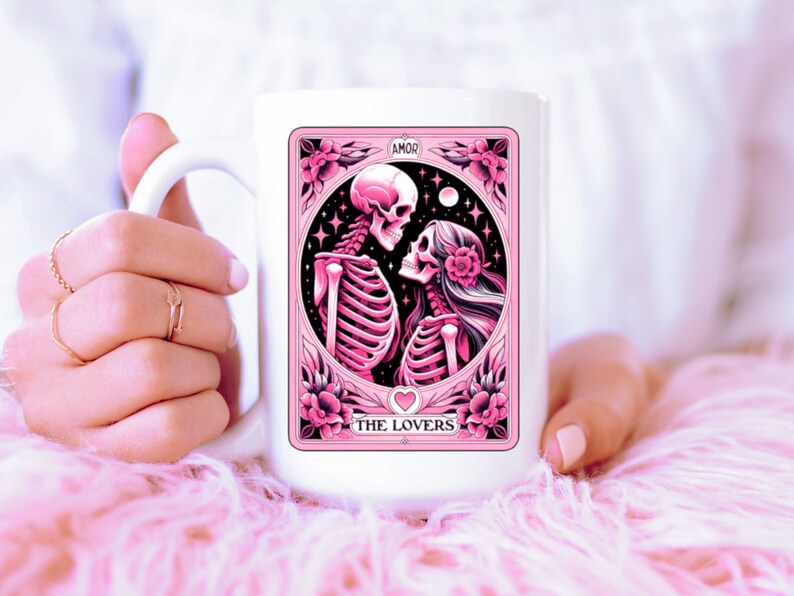 K&ouml;nnte beinhalten: Wei&szlig;e Keramik-Tasse mit einer rosa-schwarzen Illustration von zwei sich umarmenden Skeletten, mit den Worten "AMOR" und "THE LOVERS". Das Design enth&auml;lt florale und sternf&ouml;rmige Elemente.