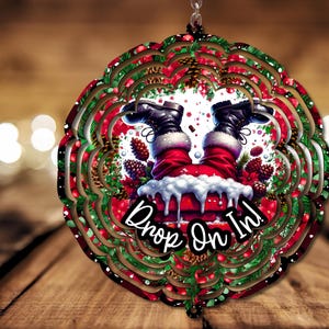 May include: A red and green Christmas wind spinner with a white background. Santa's legs are sticking out of a chimney with the text "Drop On In!"
