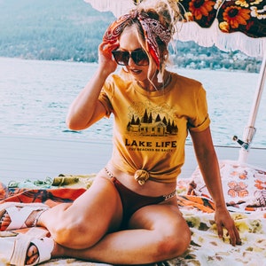May include: Mustard yellow t-shirt with the text "LAKE LIFE CUZ BEACHES BE SALTY" and a cabin graphic. The person is wearing sunglasses and a red bandana. The person is sitting on a colorful blanket near a lake.