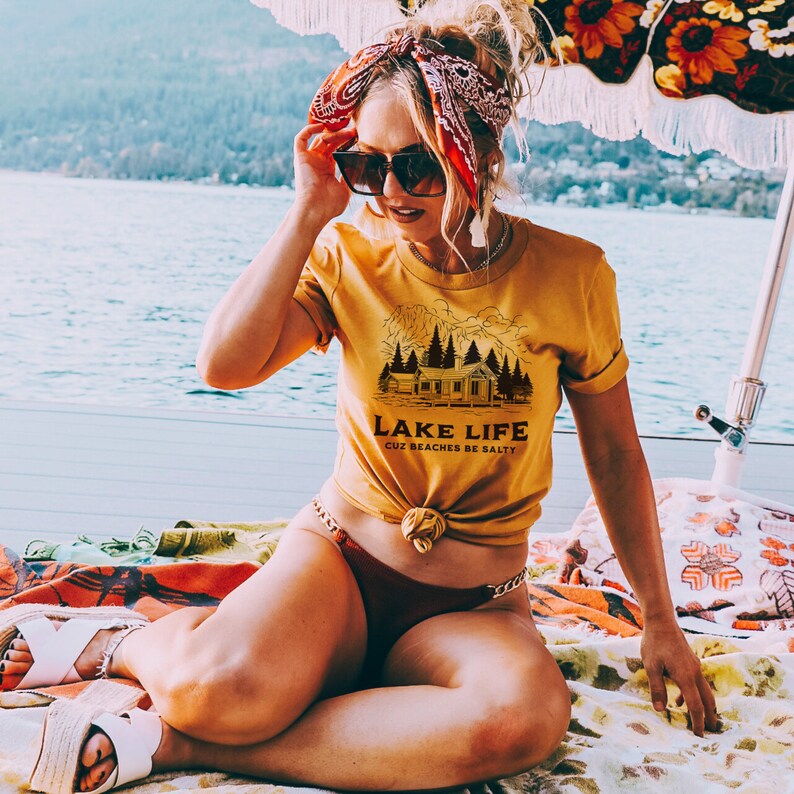 May include: Mustard yellow t-shirt with the text "LAKE LIFE CUZ BEACHES BE SALTY" and a cabin graphic. The person is wearing sunglasses and a red bandana. The person is sitting on a colorful blanket near a lake.