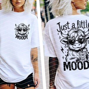 May include: White t-shirt with a black and white graphic of a cow wearing sunglasses and holding a drink. The text "Just a little MOODY" is printed on the shirt.