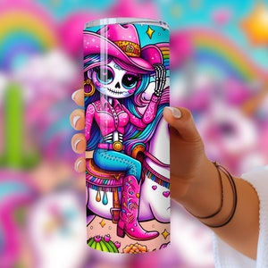 Puede incluir: Un vaso de acero inoxidable rosa y azul con una ilustraci&oacute;n colorida de un esqueleto con un sombrero de vaquero rosa montando un unicornio. El unicornio tiene una crin y una cola de arco&iacute;ris.