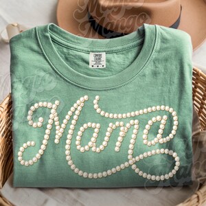 May include: A sage green t-shirt with the word "Mama" spelled out in a cursive font, using small, round, white embellishments. The shirt is folded and placed in a woven basket, with a brown hat visible in the background.